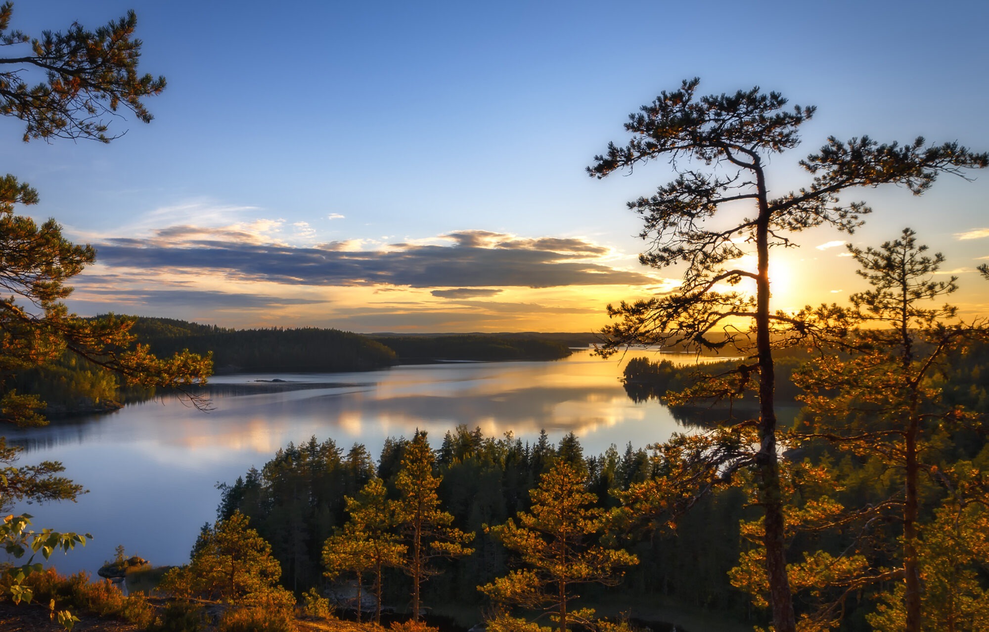 Auringonlasku Pisamalahden Linnavuorelta, metsäinen Saimaa-järvimaisema etualalla ja taivas hehkuu oranssin ja sinisen sävyissä.