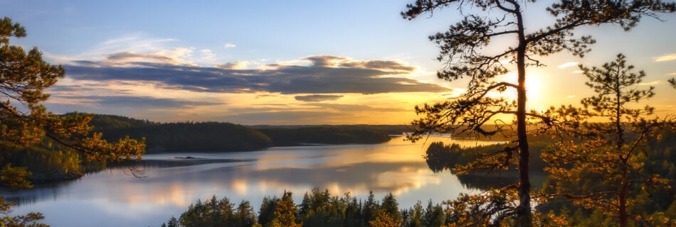 Auringonlasku Pisamalahden Linnavuorelta, metsäinen Saimaa-järvimaisema etualalla ja taivas hehkuu oranssin ja sinisen sävyissä.
